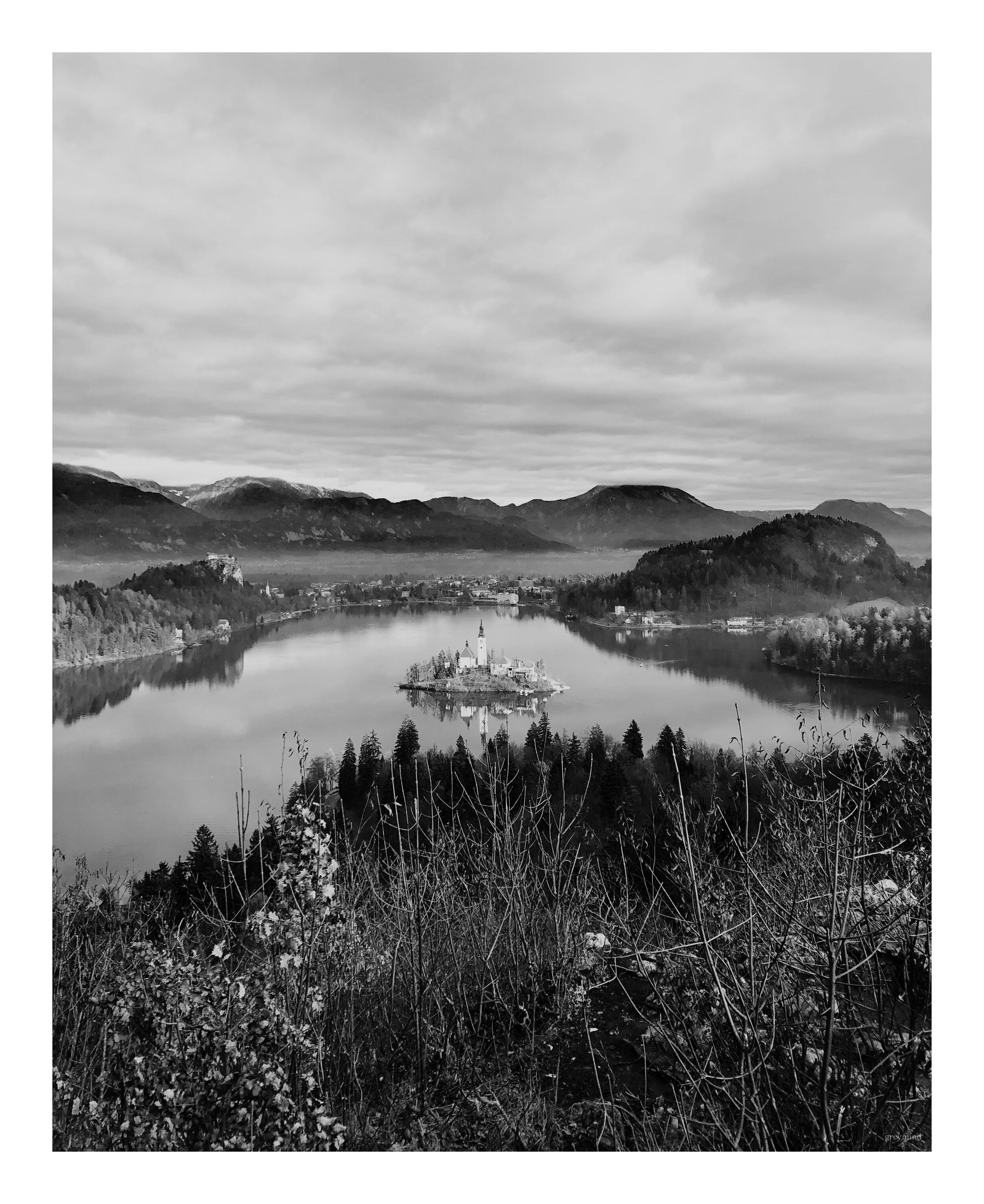Black-and-white view down onto Lake Bled and its island church, with misty hills beyond.
