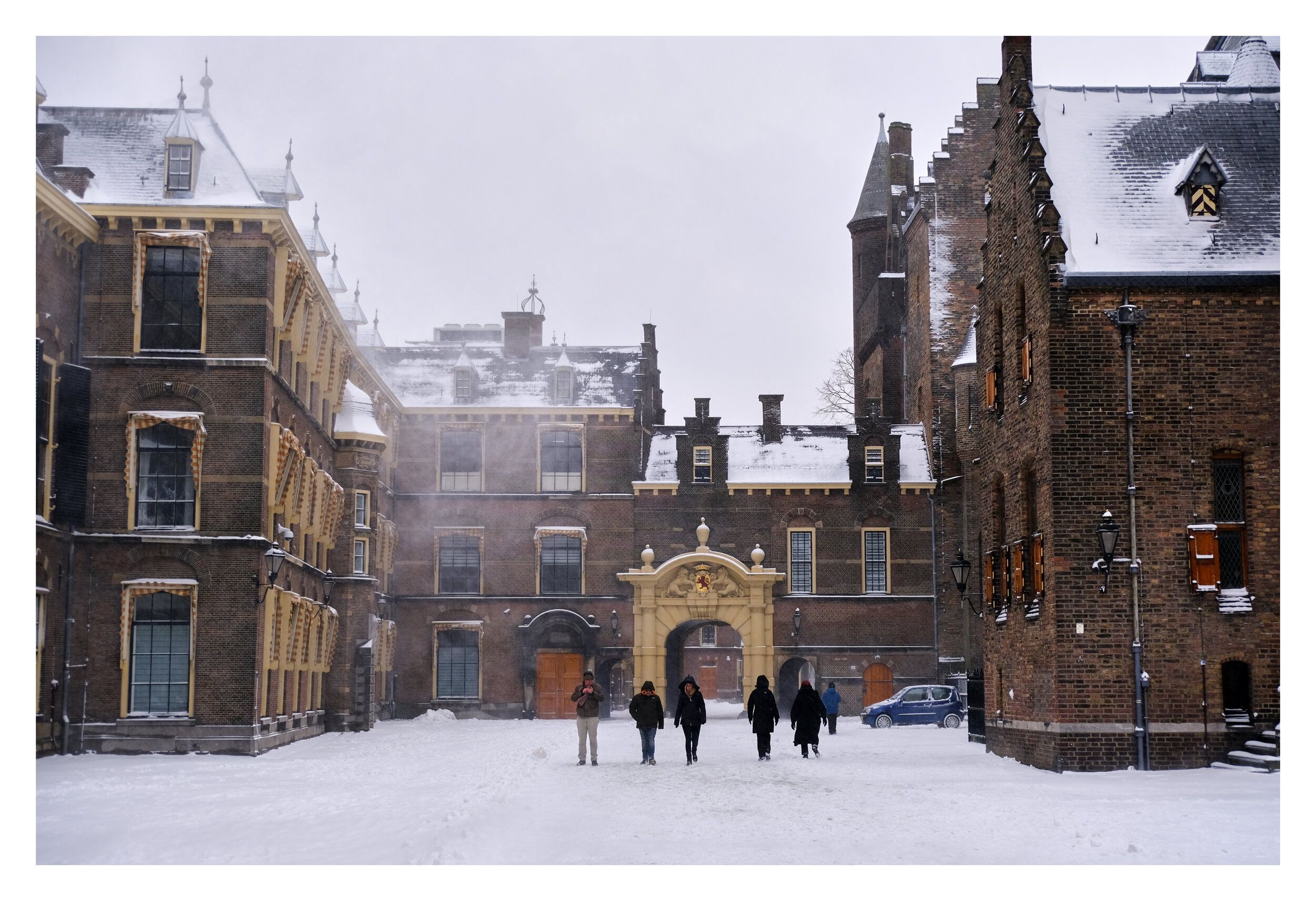 Historic brick buildings around a snowy courtyard, with people walking towards a gilded gateway.