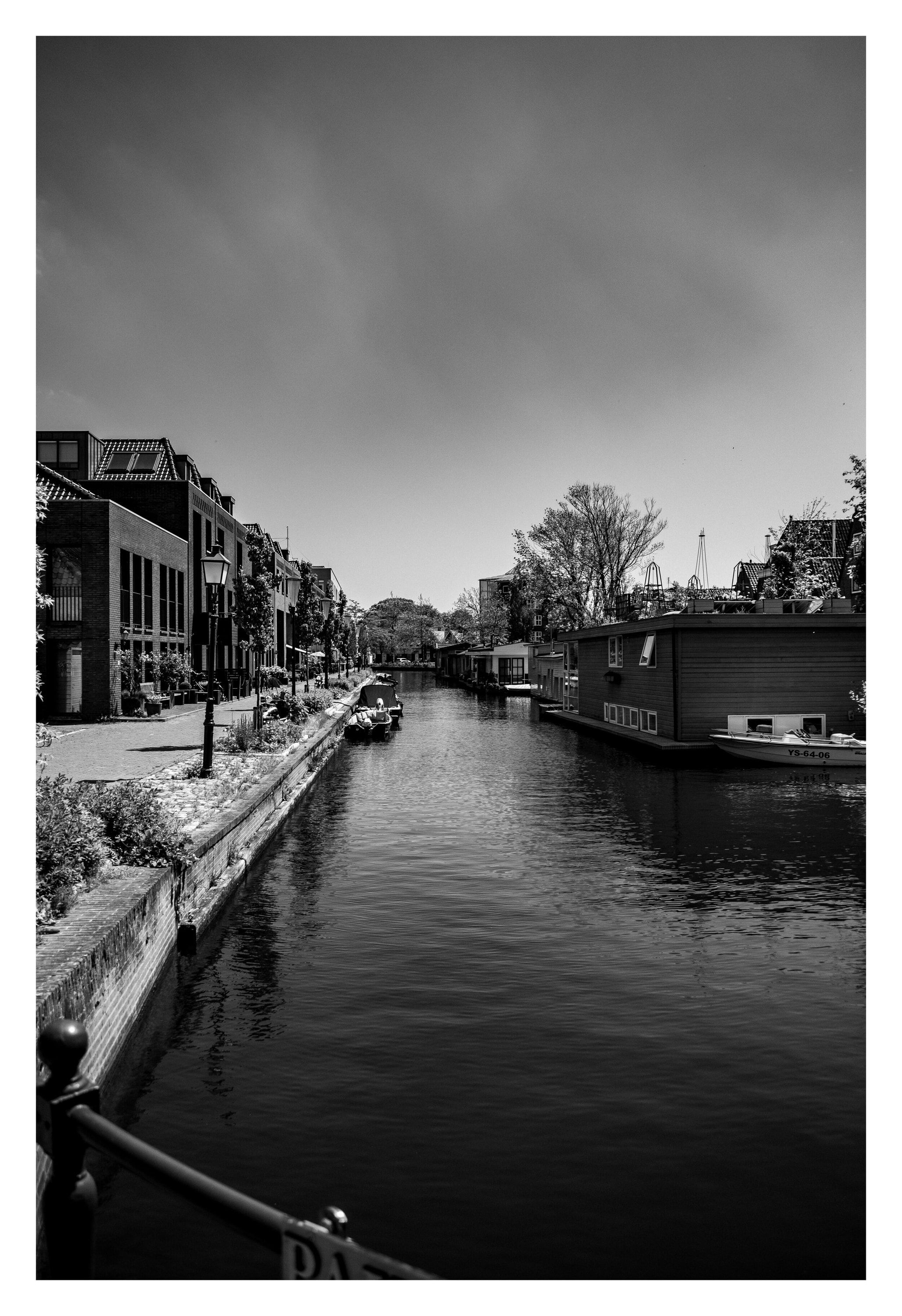 Black-and-white canal with moored boats and houses along the quay under a cloudy sky.